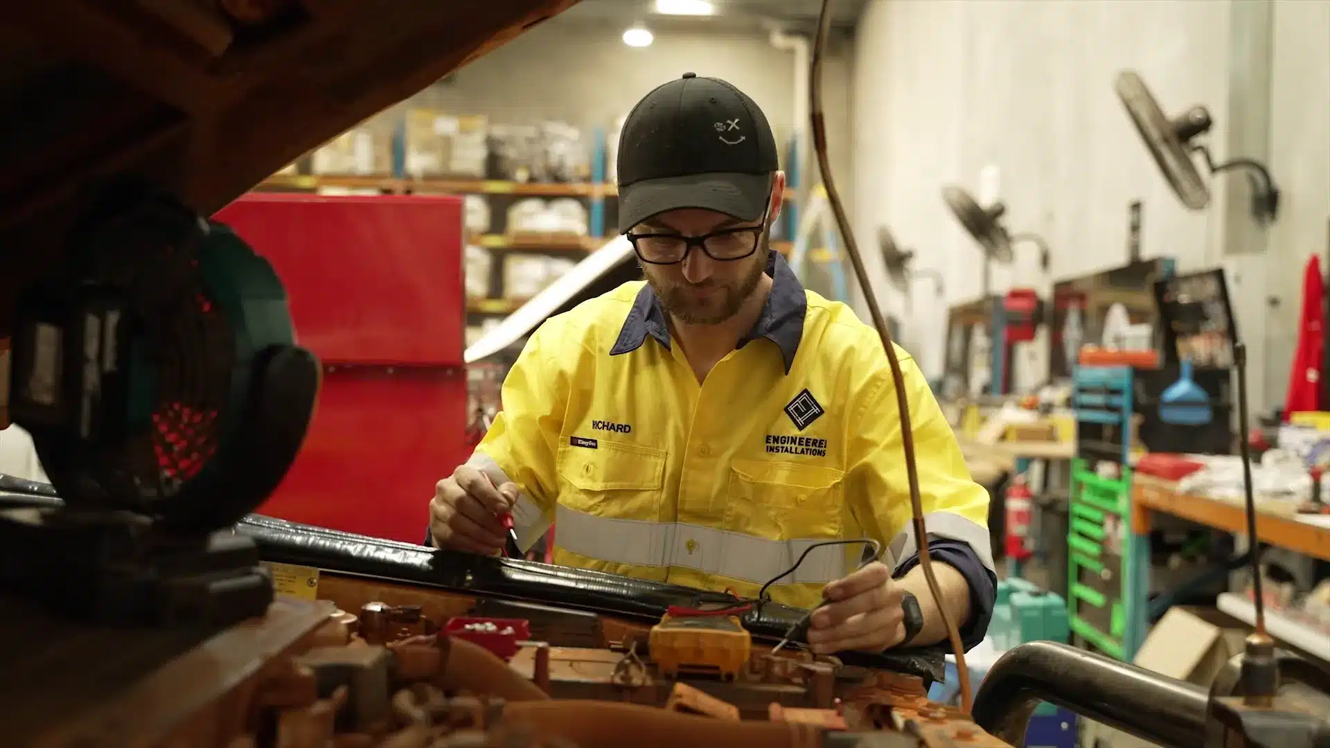 68ca650ab428a786c4f6aaf4_1746156717113 A mechanic in a yellow high-vis shirt and black cap uses a multimeter to test a vehicle engine in a workshop.